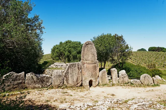 La stele centinata di Coddu Vecchiu svetta al centro dell'esedra di lastre, cielo azzurro e macchia mediterranea
